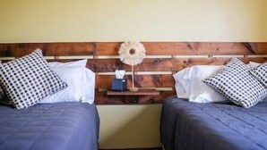 Habitación con dos camas, cabecera de madera, almohadas blancas y estampadas, una mesita de noche con lámpara y una pequeña mesita auxiliar.