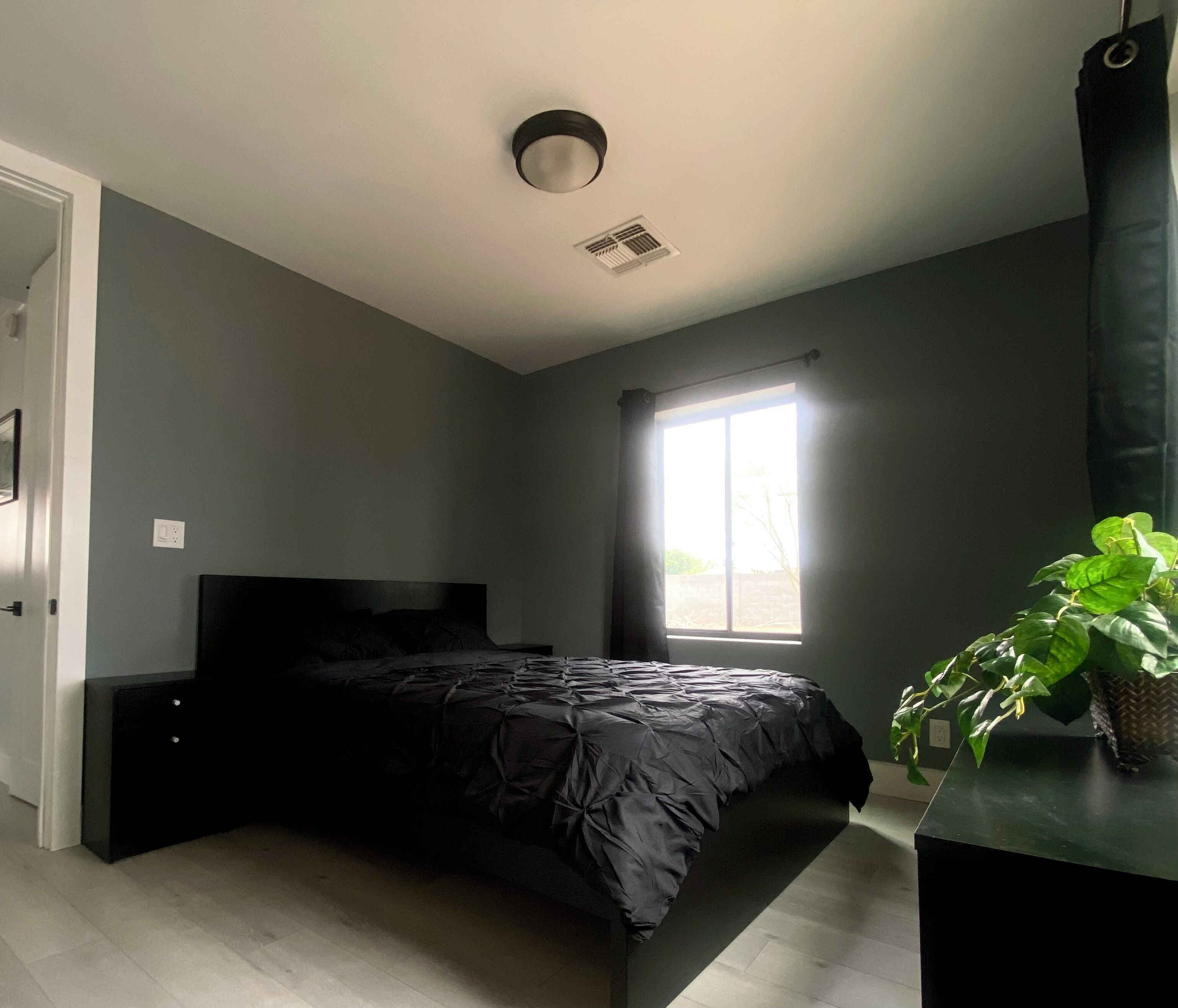 A bedroom with a bed, a window, a ceiling vent, and a potted plant.