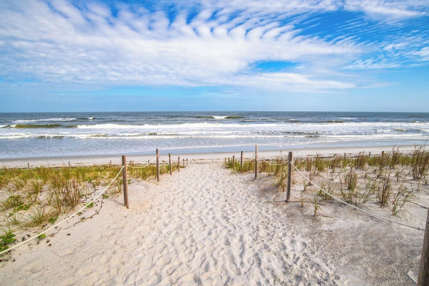 Une plage de sable bordée d’une clôture en bois, avec de l’herbe au premier plan, qui s’étend jusqu’à l’océan.