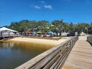 A wooden pier leading to a sandy beach area with picnic tables and umbrellas.