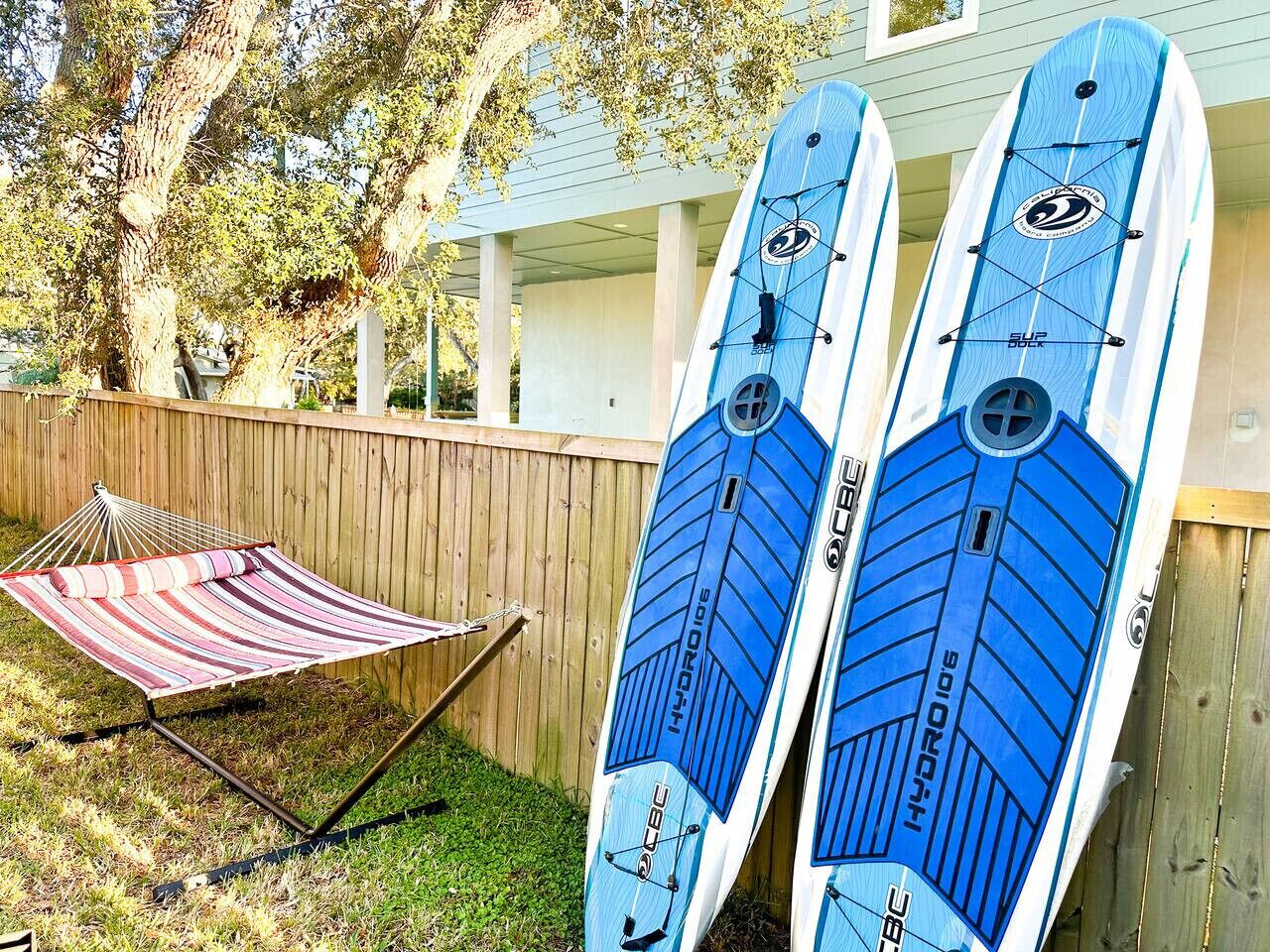 Deux planches de surf à hydrofoil appuyées contre une clôture en bois dans un jardin, avec un hamac et une maison en arrière-plan.