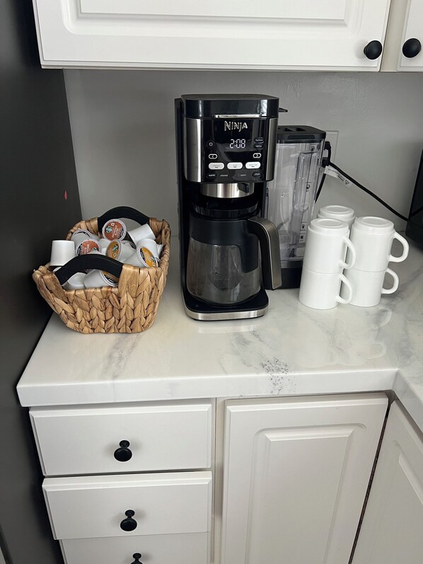Eine Kaffeemaschine, ein Korb mit Kaffeekapseln und drei weiße Tassen auf einer Küchenarbeitsfläche.