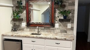 Une cuisine avec des armoires blanches, un comptoir en granit, un lave-vaisselle intégré et un miroir décoratif agrémenté d’un pare-feu en pierre.