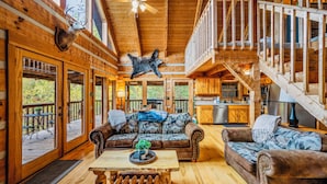 A rustic cabin interior with a wooden ceiling, a central seating area with a sofa and armchairs, a dining table, and a staircase leading to an upper level. - Stairway to Heaven •Breathtaking Mountain Views• (Sevierville)