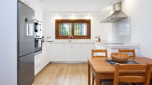 Una cocina moderna con armarios blancos, un refrigerador de acero inoxidable, un ventanal de madera sobre el fregadero y una mesa de comedor con sillas.