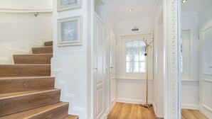 Eine Treppe mit Holzbodenstufen führt zu einer Tür, an der Wand hängt ein gerahmtes Bild und ein Fenster bietet Blick auf Bäume.