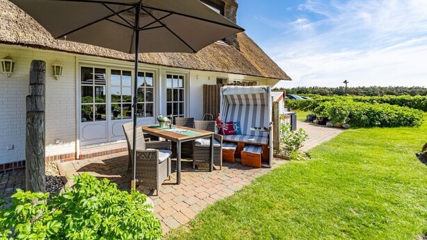 Ein Haus mit einem Reetdach und einer Terrasse, ausgestattet mit einem Tisch, Stühlen und einer gestreiften Liege.