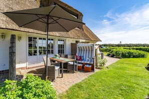 A thatched-roof house with a patio area featuring a table, chairs, and a striped lounge chair. - Adis Meerblick - Neu ab 2025 bei uns in der Vermittlung (Wenningstedt-Braderup (Sylt))