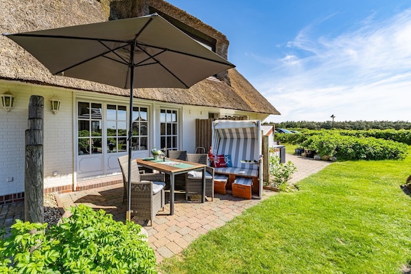 A thatched-roof house with a patio area featuring a table, chairs, and a striped lounge chair.