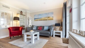 Ein Wohnzimmer mit einem roten Sessel, einem grauen Sofa und einem weißen Couchtisch. Ein Bücherregal, eine Uhr und ein Gemälde sind an der Wand zu sehen.