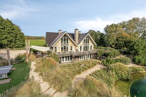 Une grande maison à deux étages, dotée d’une façade en pierre, de grandes fenêtres et d’une terrasse qui s’étend au-dessus d’un jardin aménagé.