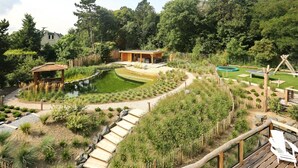 Un jardín ajardinado con un estanque, terraza de madera y una pequeña construcción.