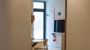 Ein Hotelzimmer mit einem Bett, einem Sessel und einem Tisch. An der Wand hängt ein Feuerlöscher, und eine Tür führt zur Küche.