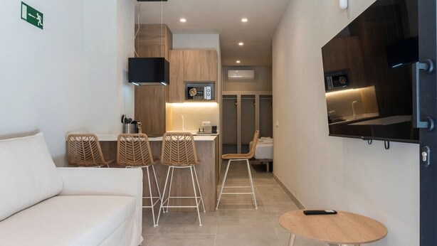 Una habitación de hotel moderna con sofá, dos taburetes de bar, una pequeña mesa redonda, un televisor de pantalla plana montado en la pared y una kitchenette con microondas y refrigerador pequeño.