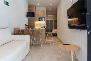A modern hotel room with a sofa, two bar stools, a small round table, a flat-screen TV mounted on the wall, and a kitchenette area with a microwave and a small refrigerator. - VLC HOST - Palau D (Valencia)