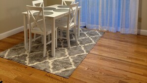 Un espace repas comprenant une table et des chaises, un tapis à motifs, un plancher en bois, un luminaire au plafond et une fenêtre ornée de rideaux légers.