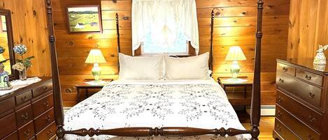 Una cama con dosel, con colcha estampada, cabecera y piecera de madera, flanqueadas por mesitas de noche con lámparas y un aparador con cajones.