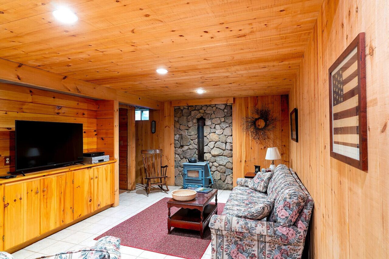 A cozy wooden cabin interior with a stone fireplace, a television on a wooden cabinet, and a floral sofa.