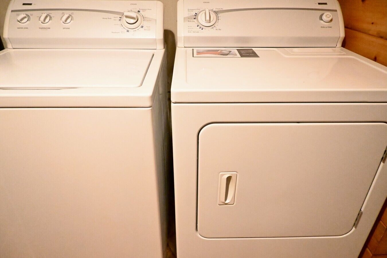 Two white front-loading washing machines with control panels, a door handle, and a label on the right machine.