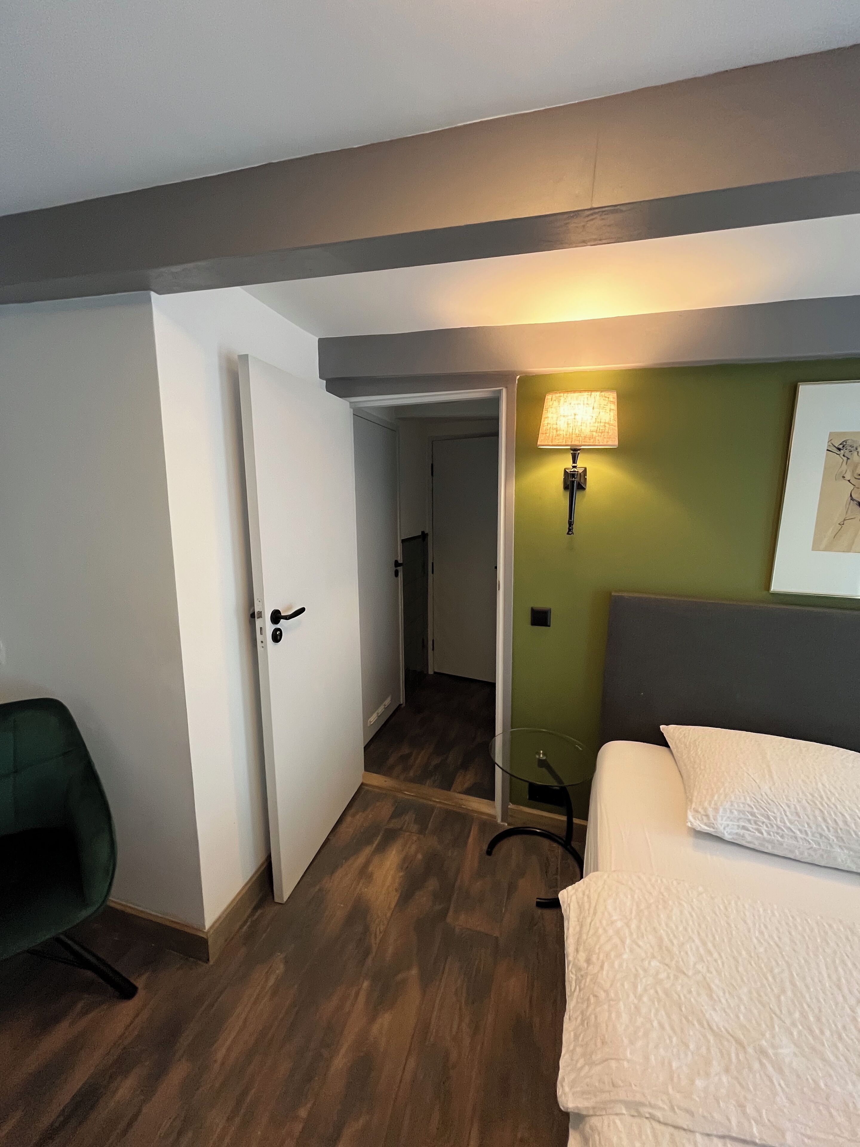 Ein Hotelzimmer mit Bett, Sessel, Schreibtisch, Lampe und einer Tür zu einem Flur.