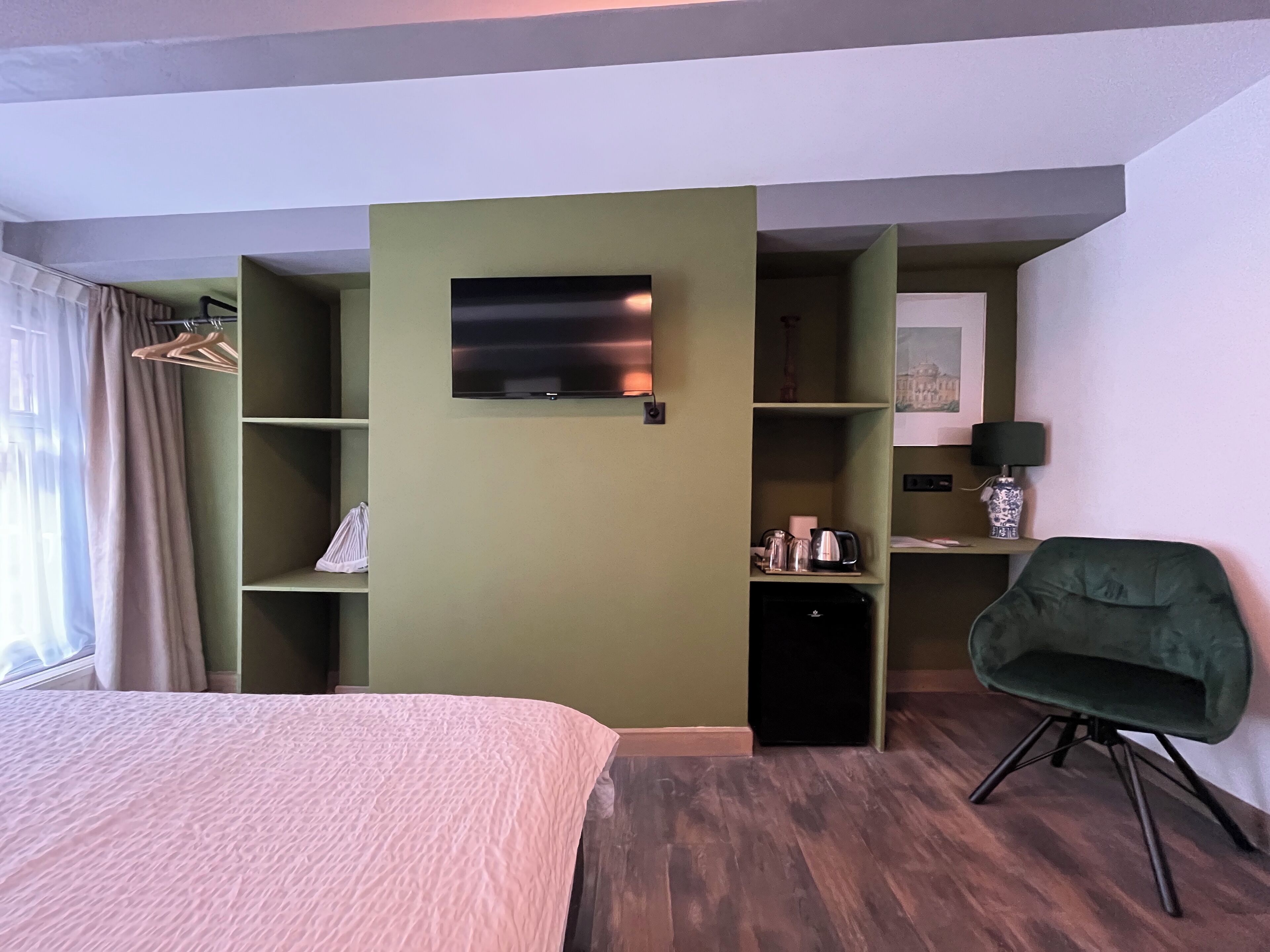 A hotel room with a bed, a desk with a computer, a chair, a TV mounted on the wall, and shelves with various items.
