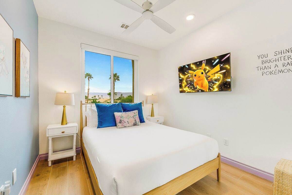 Quarto com cama, duas mesinhas de cabeceira, ventilador de teto, janela com vista e uma obra de arte na parede.