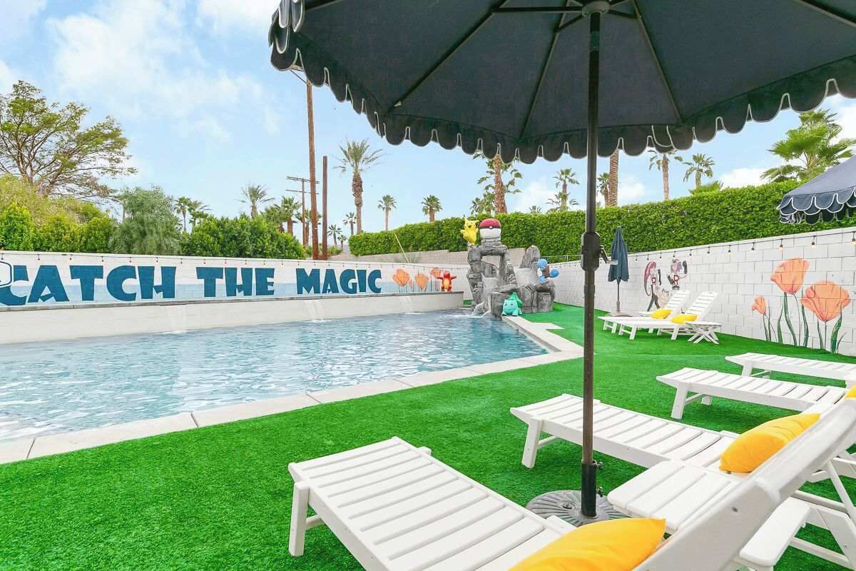 Une piscine, avec une pancarte indiquant « Laissez-vous envoûter », entourée de chaises longues blanches dotées de coussins jaunes, d’une statue de pierre et d’une fresque murale.