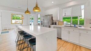 A modern kitchen with white cabinetry, a large island with bar stools, stainless steel appliances, and pendant lighting. - Catch The Magic- Pokémon Slide, Arcade, Pickleball (Indio)