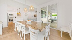 A modern dining area with a glass-top table, white chairs, pendant lights, and a view into the kitchen. - Catch The Magic- Pokémon Slide, Arcade, Pickleball (Indio)