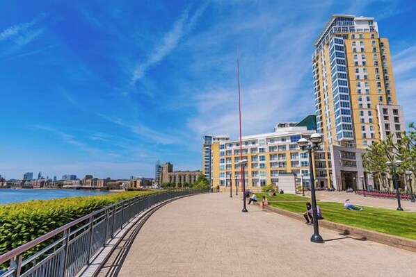 A waterfront promenade with a high-rise building, a clear sky, and a calm river.
