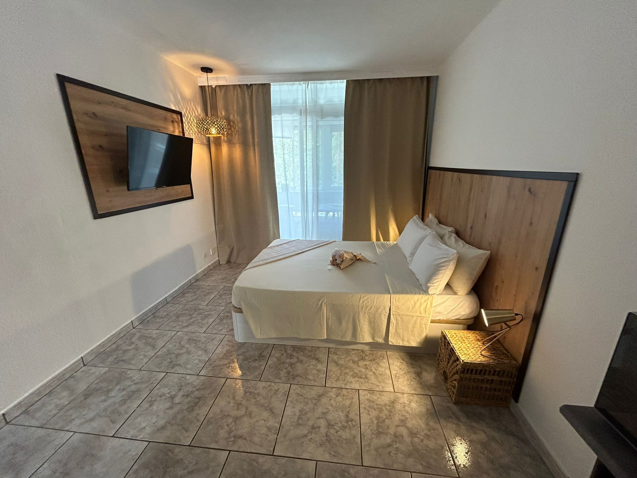 A hotel room with a bed, a TV mounted on the wall, a window with curtains, and a small wicker basket.