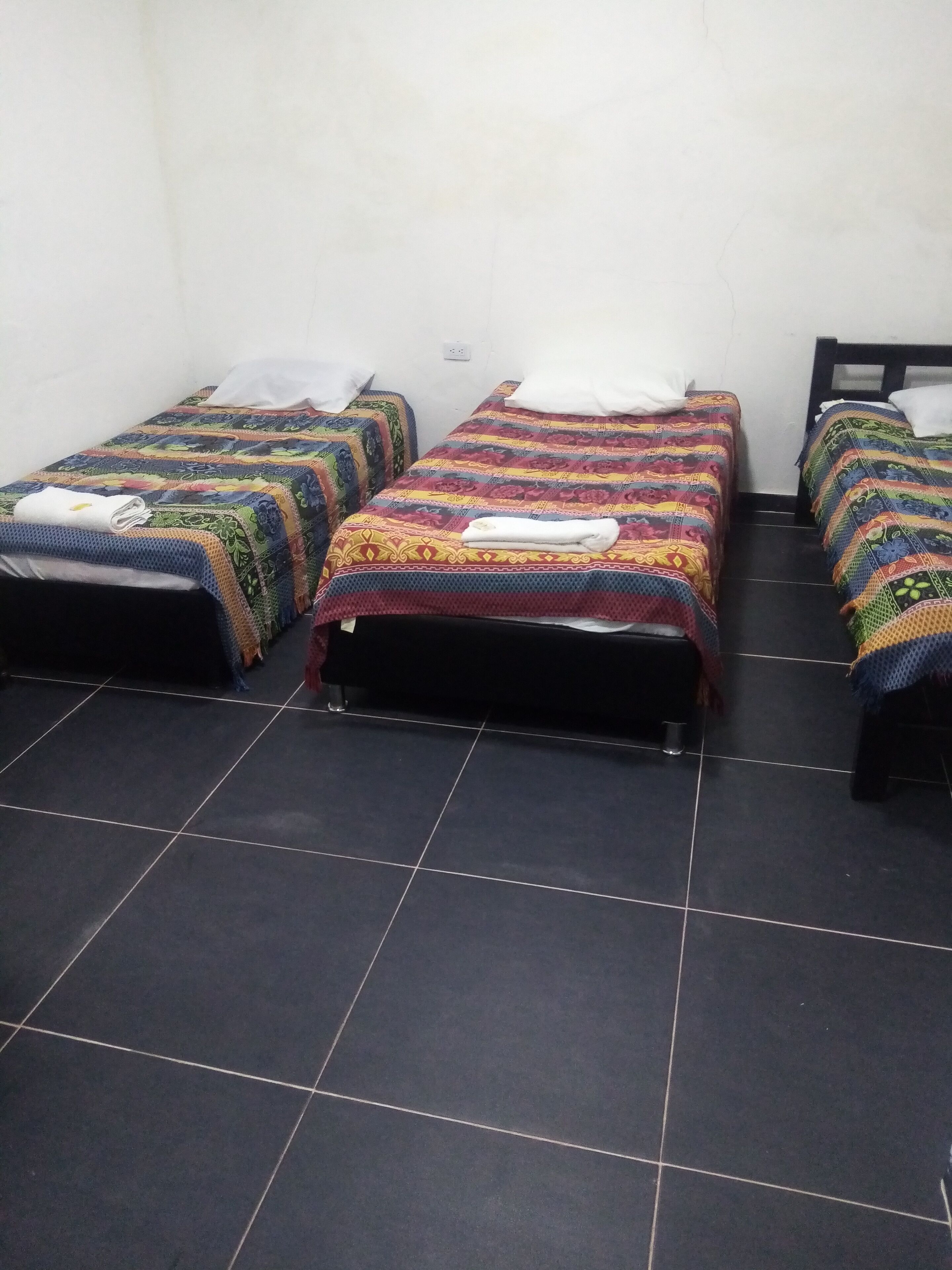 A room with three single beds, each with a colorful quilt and a white pillow. The room has a tiled floor and plain white walls.