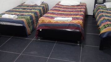 Habitación con tres camas individuales, cada una con una colcha de colores y un almohadón blanco. El piso es de baldosas y las paredes son lisas y de color blanco.