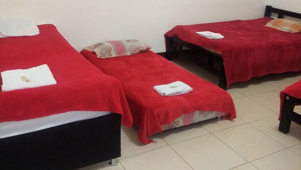 A room with three single beds, each with a red bedspread and a white towel. The room has white walls and a tiled floor.