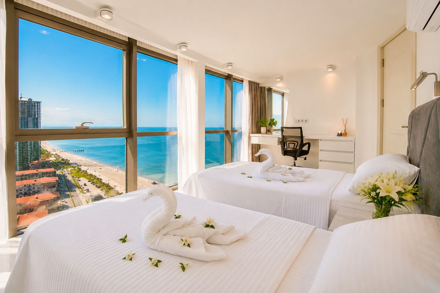 A hotel room with two beds, a desk, a chair, and a large window with a view of the beach and ocean.