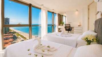 A hotel room with two beds, a desk, a chair, and a large window with a view of the beach and ocean.