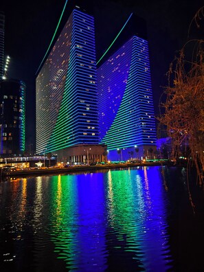 Une vue nocturne de deux gratte-ciel illuminés, dont les reflets se mirent dans une étendue d’eau.