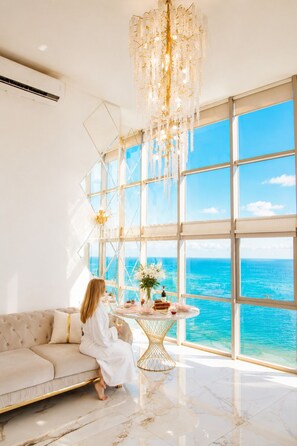 A room with a large window, a chandelier, a round table with a vase and wine bottle, and a sofa. - Panorama (Batumi)