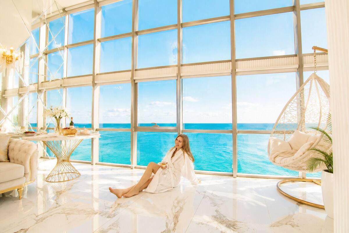 A spacious room with large windows, a marble floor, a white sofa, a hanging chair, and a table with a bottle and flowers.