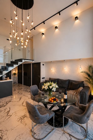 A modern living room with a marble floor, a glass coffee table, grey upholstered chairs, a sofa, and a hanging light fixture.