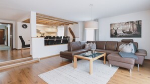 Ein modernes Wohnzimmer mit einer braunen Ledersofa, einem Glastisch und einem weißen Teppich. Ein Essbereich mit Holztisch und Stühlen ist zu sehen, ebenso eine Küche durch eine offene Tür.