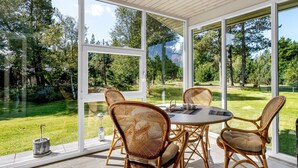 A covered patio with a round table and wicker chairs, large glass windows, and a view of a grassy area with trees.