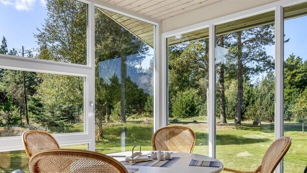 A covered patio with wicker chairs and a table, large windows offering a view of a forested area.