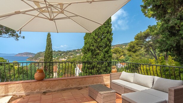 Una terraza con sombrilla blanca, mobiliario de mimbre y vistas al mar y las colinas.
