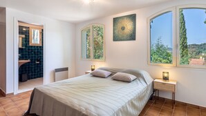 Un dormitorio con una cama, dos mesitas de noche con lámparas, un ventanal con vistas a zonas verdes y un baño con pared de azulejos azules y un lavabo.