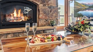 A stone fireplace with a fire, a wooden table with a charcuterie board, two glasses of water, and a television displaying a scenic landscape. - Stunning 6BR Lodge w/ Beautiful Mt. Quandary Views (Blue river)