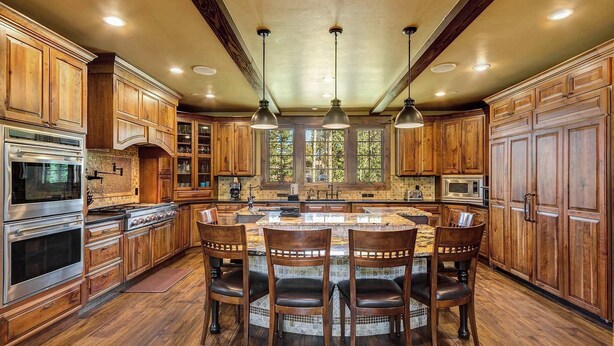 A spacious kitchen with wooden cabinets, a central island with bar stools, stainless steel appliances, and large windows.