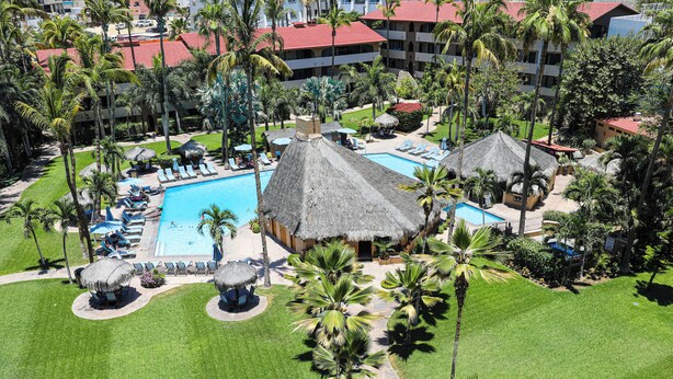 An aerial view of a resort with a central pool, thatched-roof structures, and lush landscaping. - A-609 Vacation in this Exquisite Spacious 2-bedroom condominium! (Cabo San Lucas)