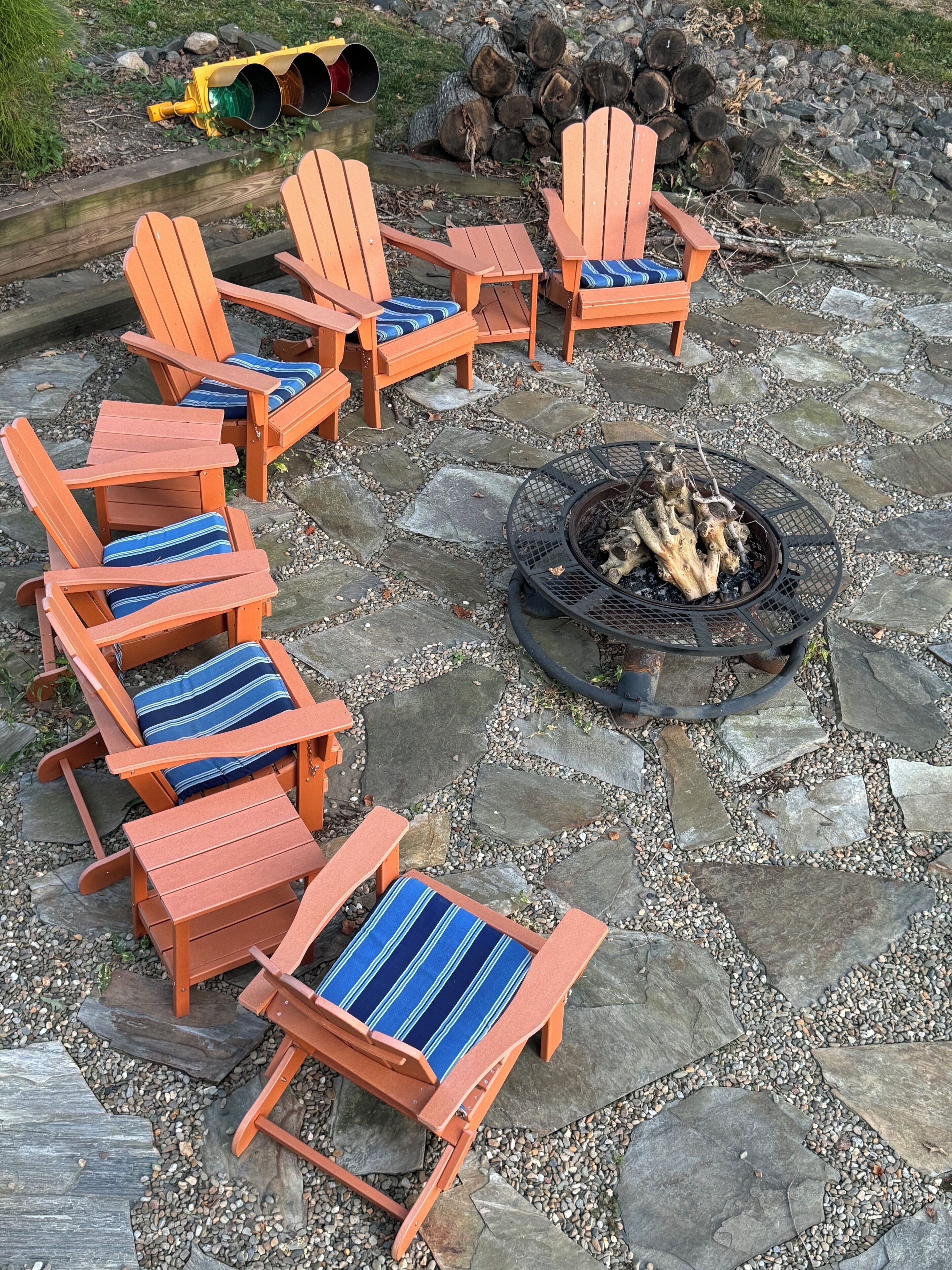 Un hogar con fuego rodeado por seis sillas Adirondack con cojines a rayas azules, dispuestas en un patio de piedra.