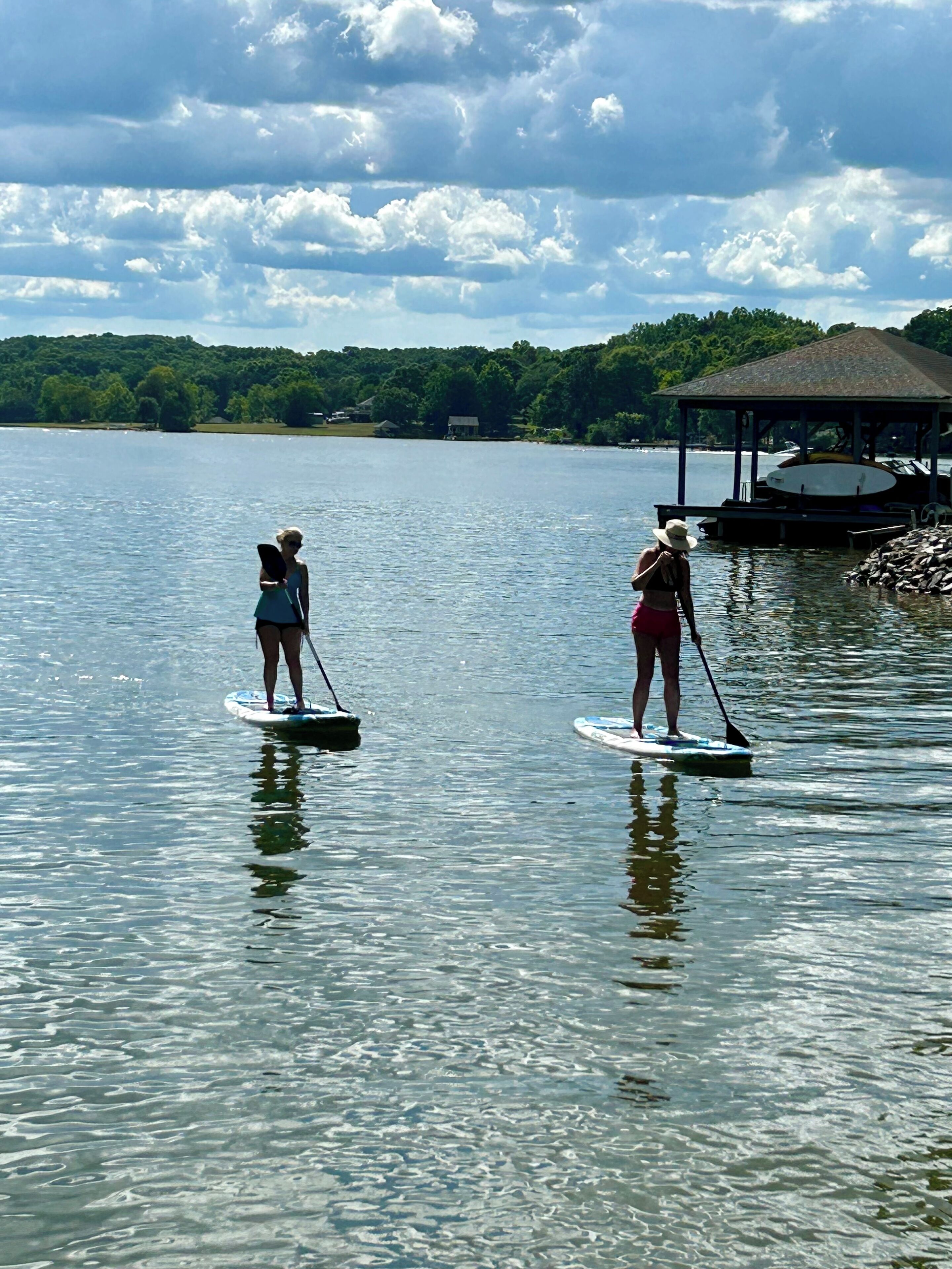 Duas pessoas praticando paddleboard em um lago calmo, com um píer e um pavilhão ao fundo.
